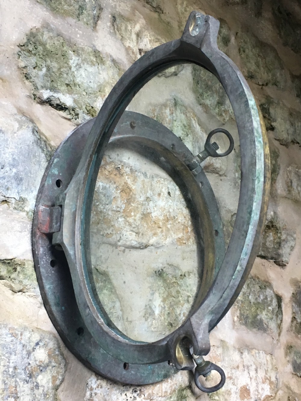 A heavy antique bronze circular ship porthole featuring visible hinge mounts, dog-bolt fasteners, a clear glass pane, and natural green patina. View XIV