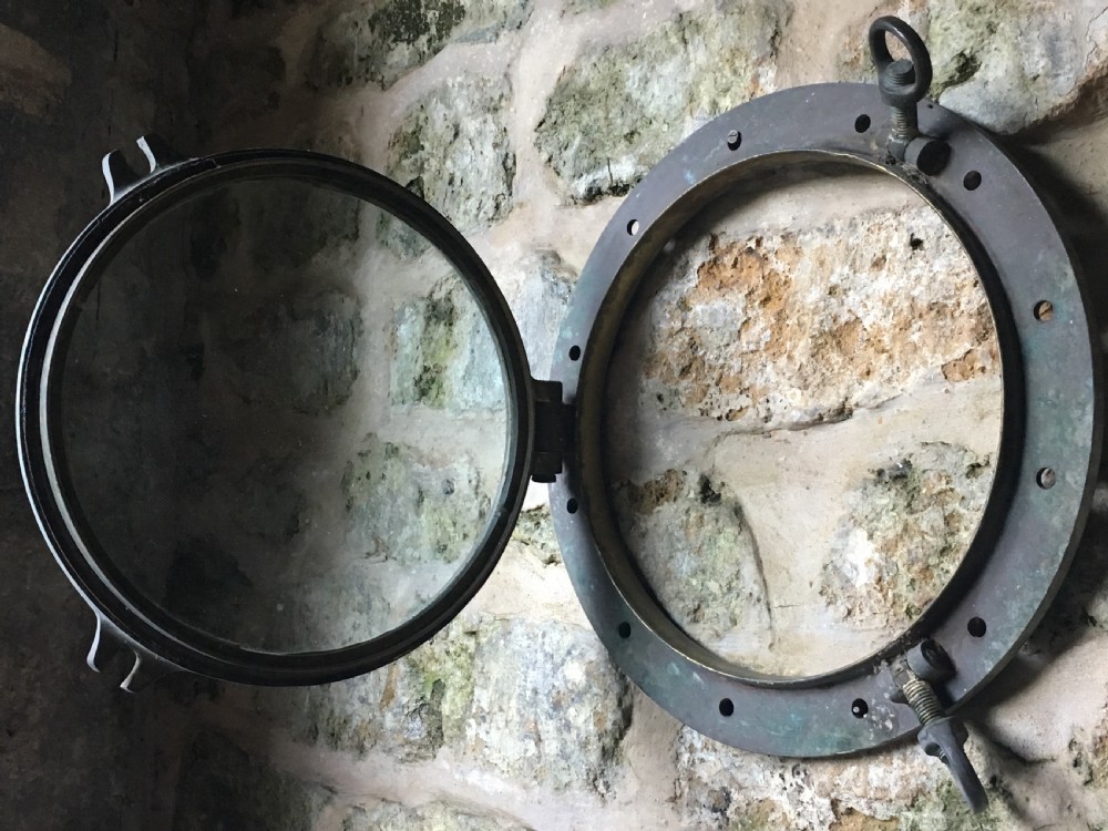A heavy antique bronze circular ship porthole featuring visible hinge mounts, dog-bolt fasteners, a clear glass pane, and natural green patina. View XV