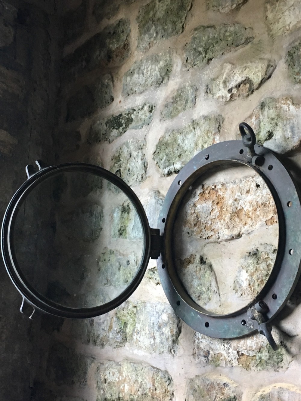 A heavy antique bronze circular ship porthole featuring visible hinge mounts, dog-bolt fasteners, a clear glass pane, and natural green patina. View XVI