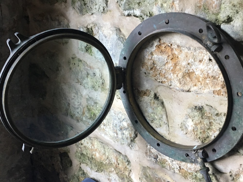 A heavy antique bronze circular ship porthole featuring visible hinge mounts, dog-bolt fasteners, a clear glass pane, and natural green patina. View XVII
