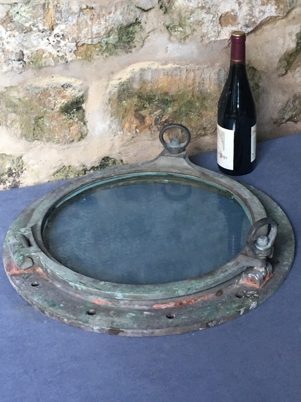 A heavy antique bronze circular ship porthole featuring visible hinge mounts, dog-bolt fasteners, a clear glass pane, and natural green patina. View XVIII