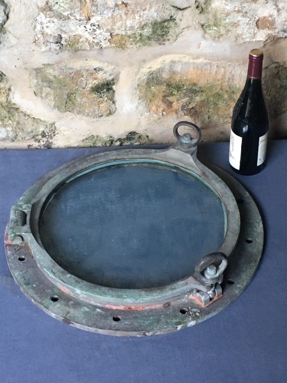 A heavy antique bronze circular ship porthole featuring visible hinge mounts, dog-bolt fasteners, a clear glass pane, and natural green patina. View XIX
