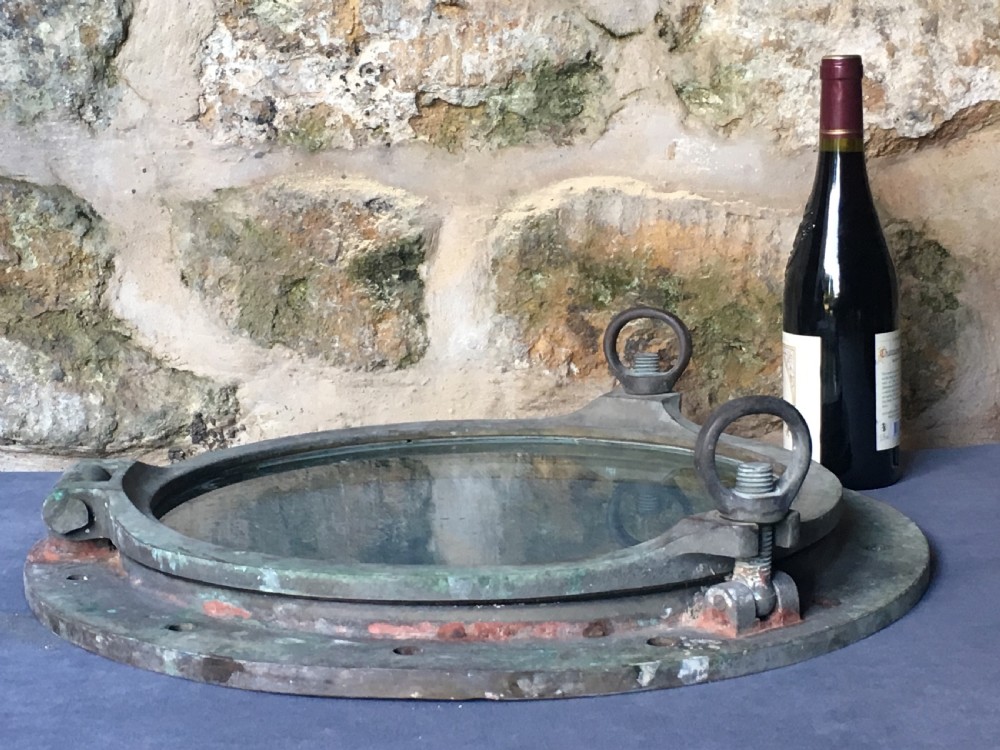 A heavy antique bronze circular ship porthole featuring visible hinge mounts, dog-bolt fasteners, a clear glass pane, and natural green patina. View XX