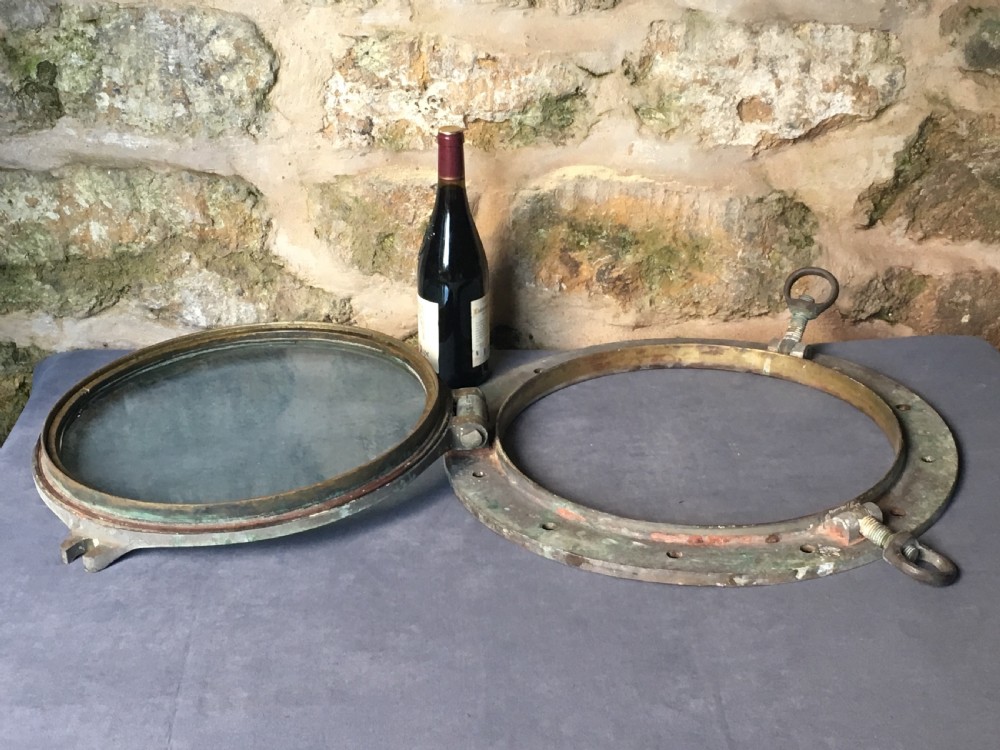 A heavy antique bronze circular ship porthole featuring visible hinge mounts, dog-bolt fasteners, a clear glass pane, and natural green patina. View XXI
