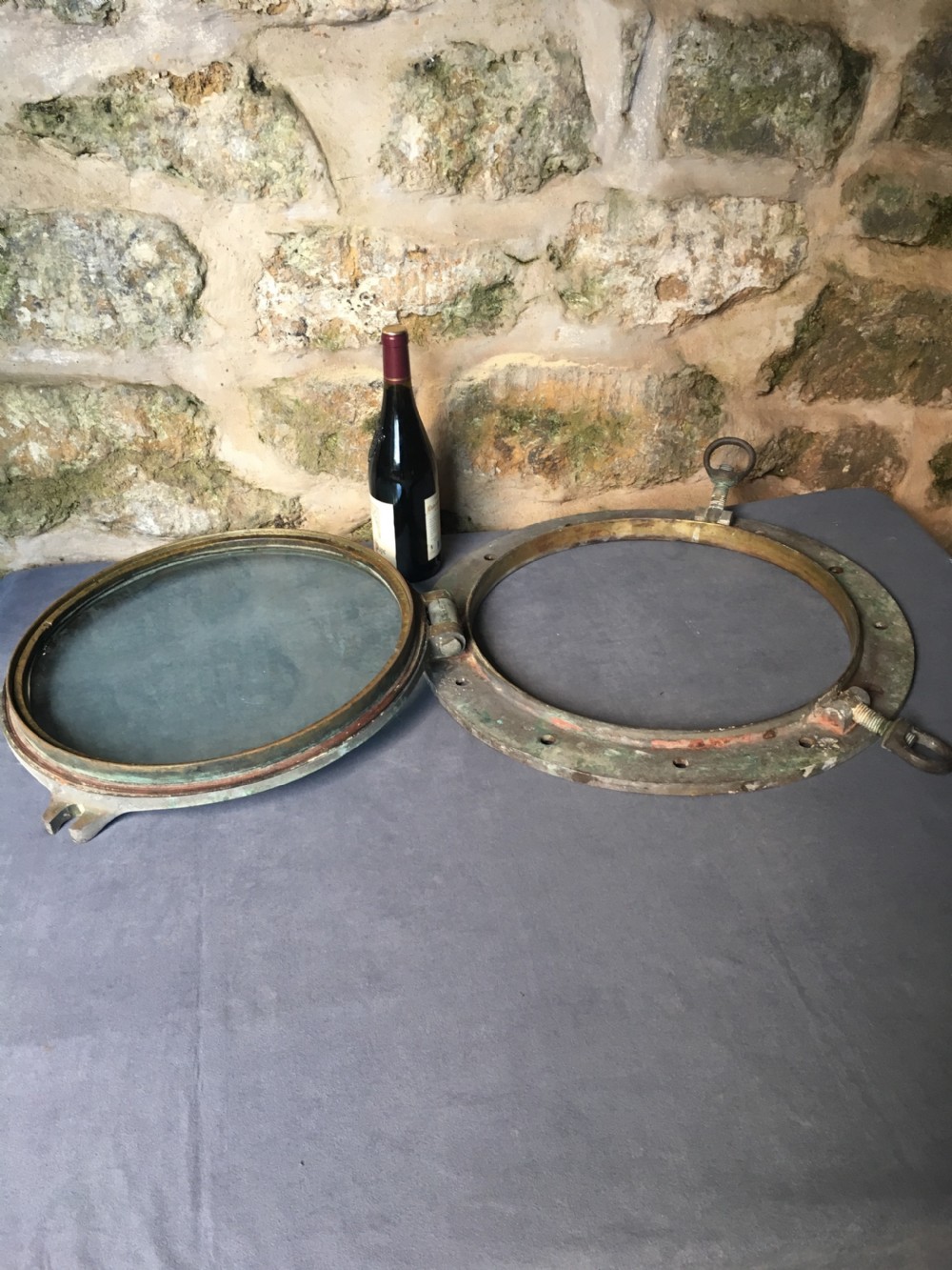 A heavy antique bronze circular ship porthole featuring visible hinge mounts, dog-bolt fasteners, a clear glass pane, and natural green patina. View XXII
