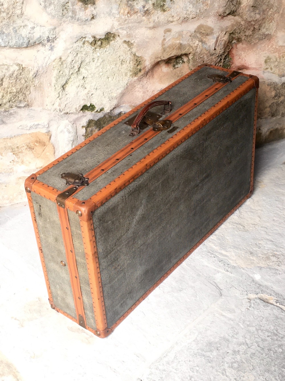 An antique French hard-sided luggage suitcase featuring grey canvas, tan leather trim, brass hardware, and a leather carrying handle.