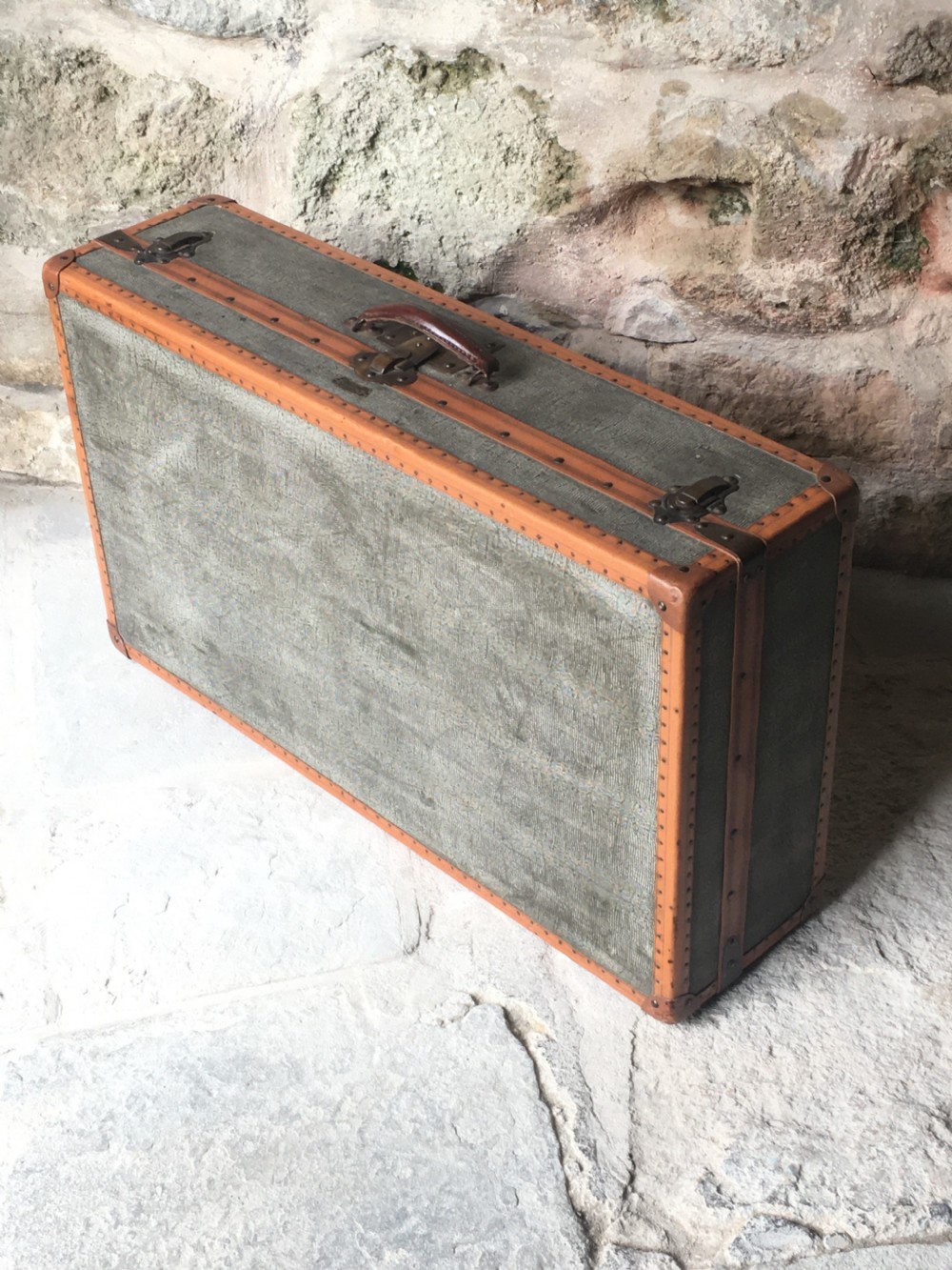 An antique French hard-sided luggage suitcase featuring grey canvas, tan leather trim, brass hardware, and a leather carrying handle. View II