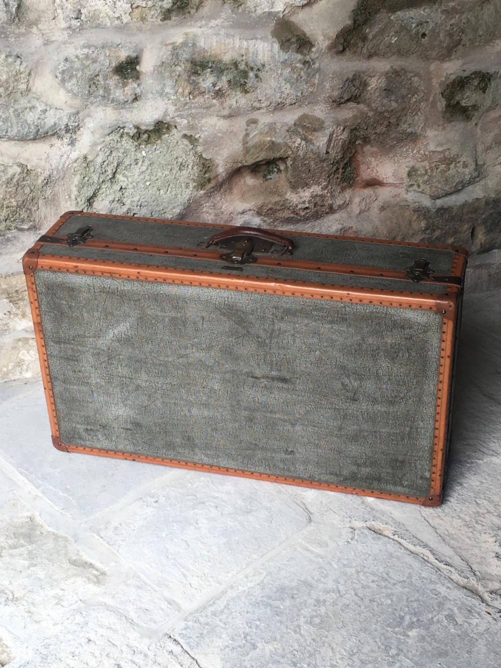 An antique French hard-sided luggage suitcase featuring grey canvas, tan leather trim, brass hardware, and a leather carrying handle. View III