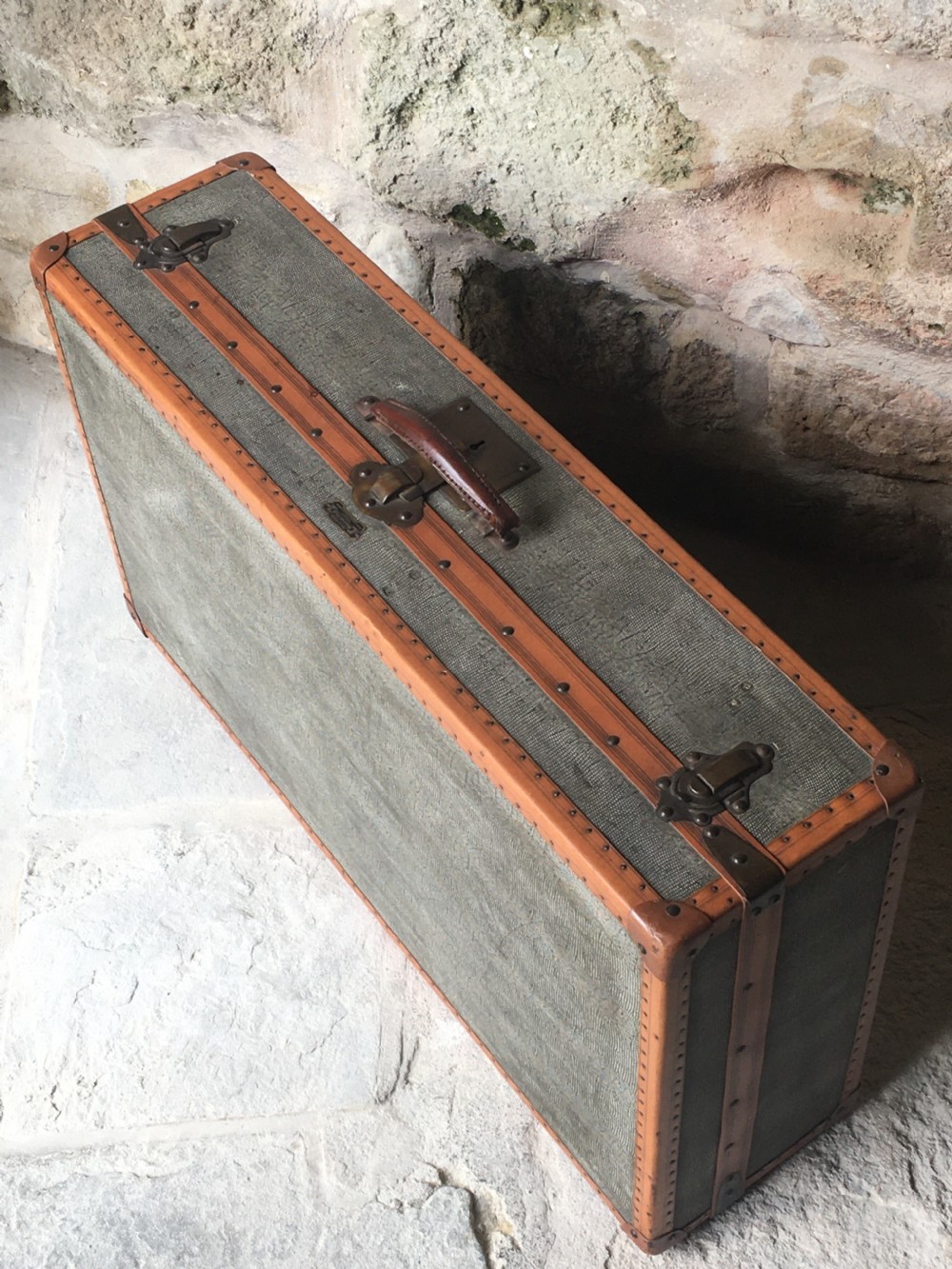 An antique French hard-sided luggage suitcase featuring grey canvas, tan leather trim, brass hardware, and a leather carrying handle. View V