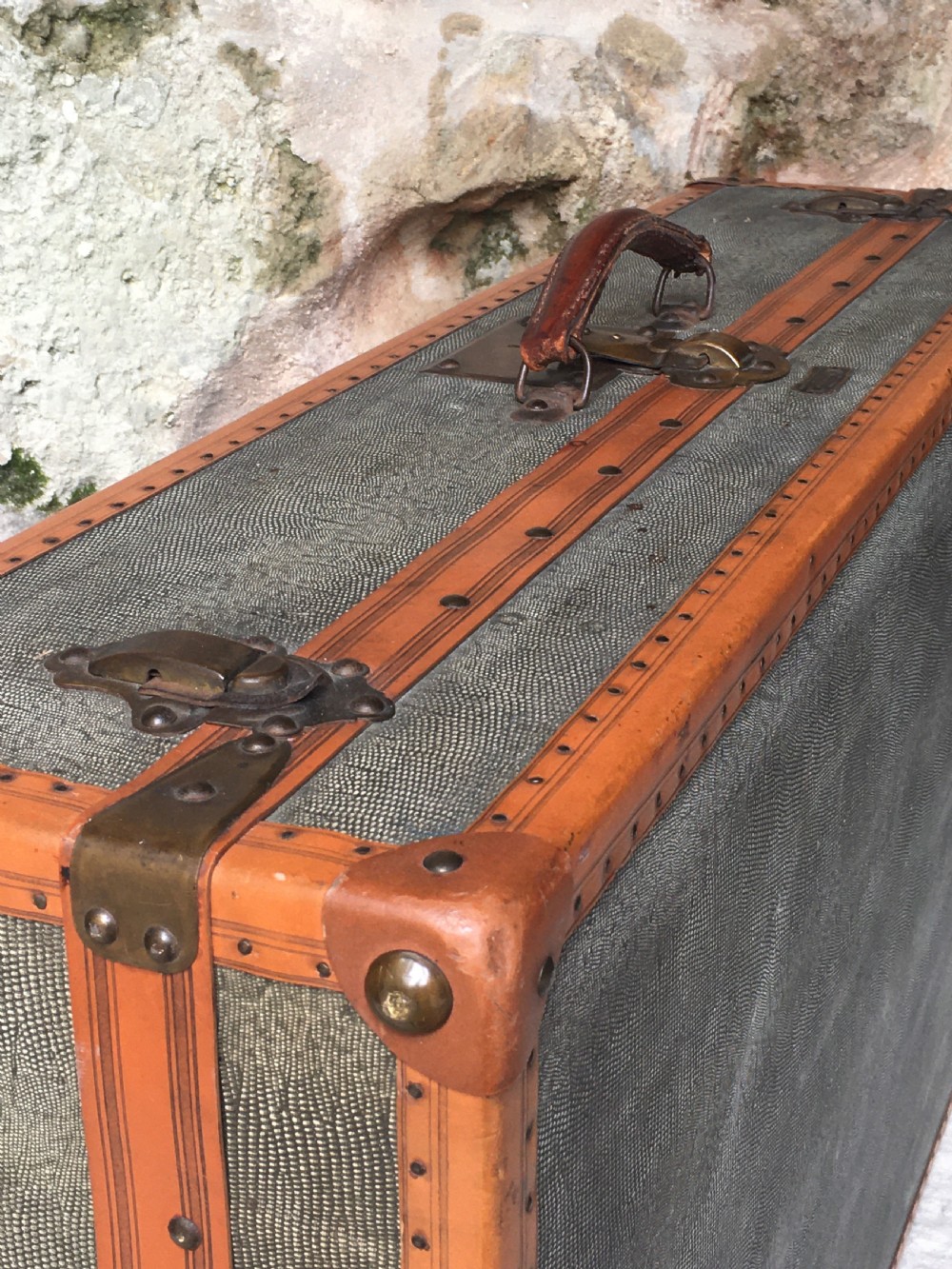 An antique French hard-sided luggage suitcase featuring grey canvas, tan leather trim, brass hardware, and a leather carrying handle. View VI