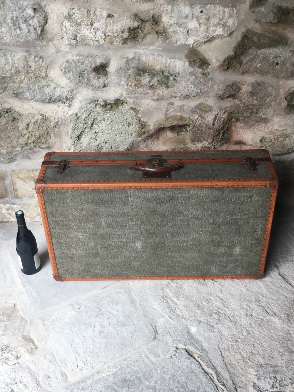 An antique French hard-sided luggage suitcase featuring grey canvas, tan leather trim, brass hardware, and a leather carrying handle. View VII