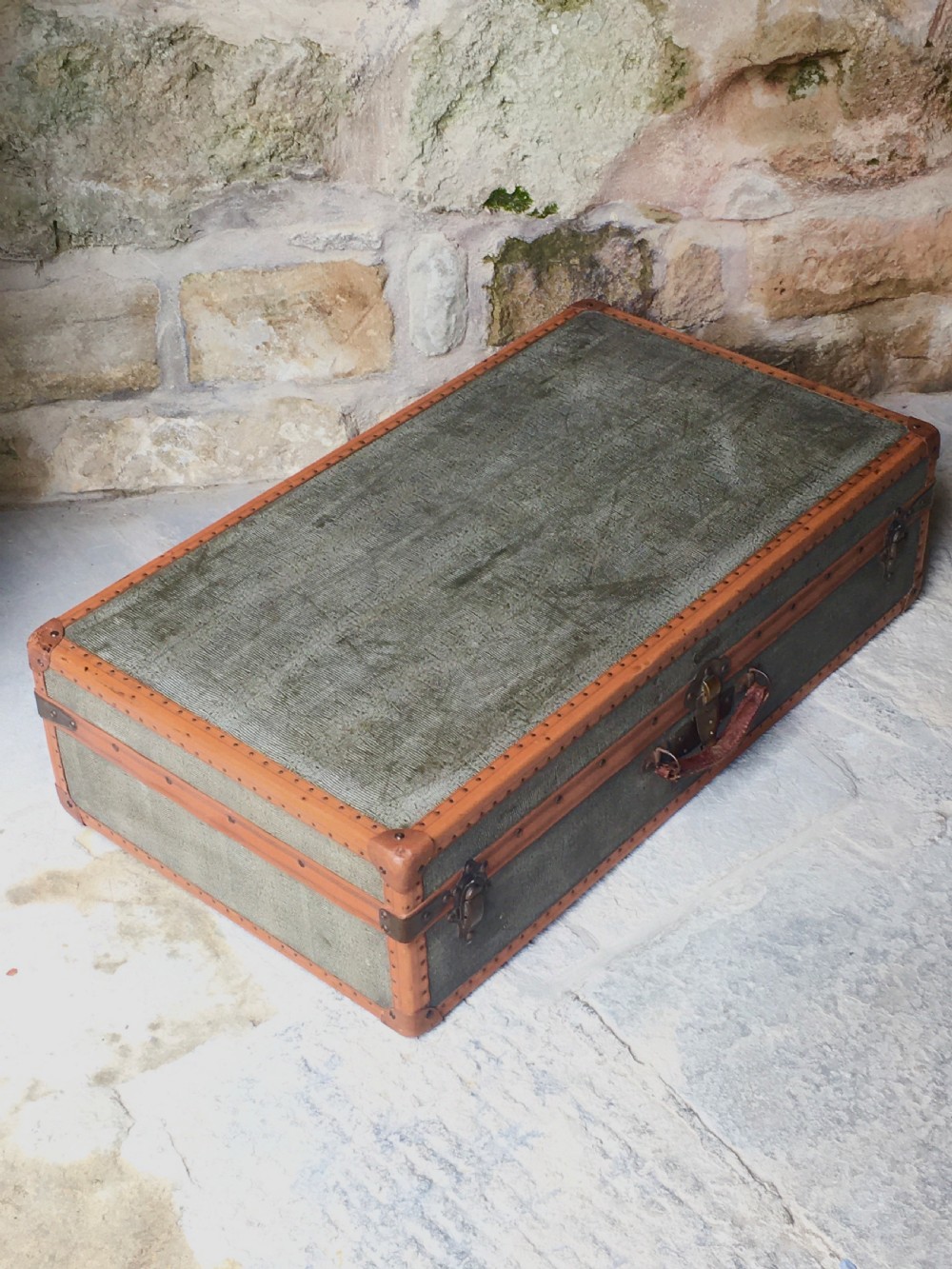 An antique French hard-sided luggage suitcase featuring grey canvas, tan leather trim, brass hardware, and a leather carrying handle. View X