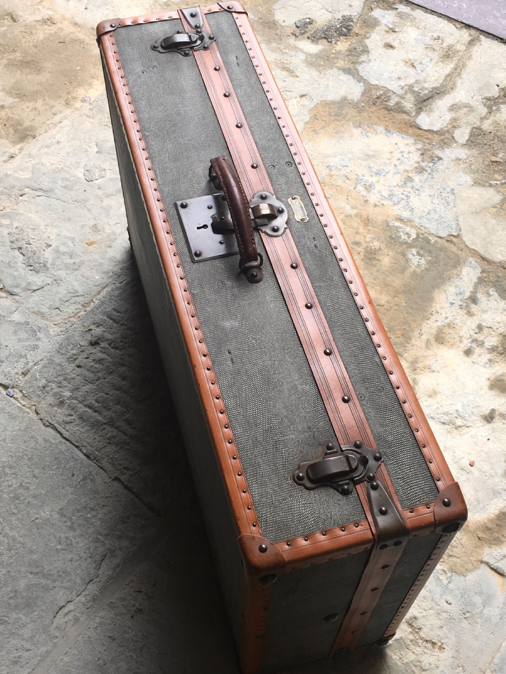 An antique French hard-sided luggage suitcase featuring grey canvas, tan leather trim, brass hardware, and a leather carrying handle. View XXIV