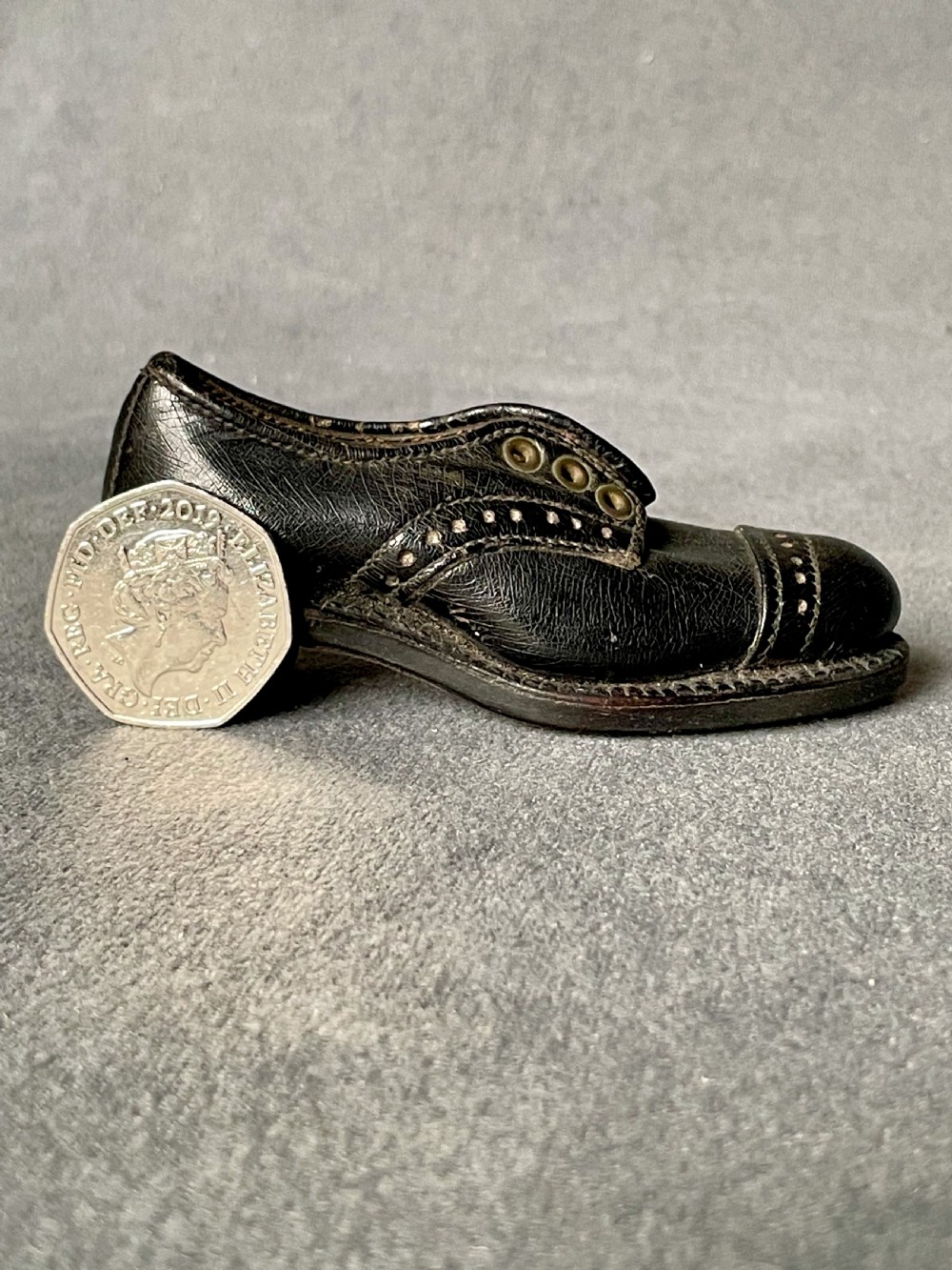A miniature black leather shoe crafted with brogue stitching and metal eyelets displayed next to a coin for scale.