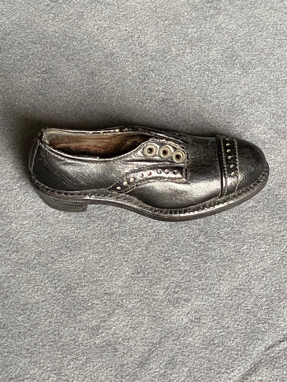 A miniature black leather shoe crafted with brogue stitching and metal eyelets displayed next to a coin for scale. View IV