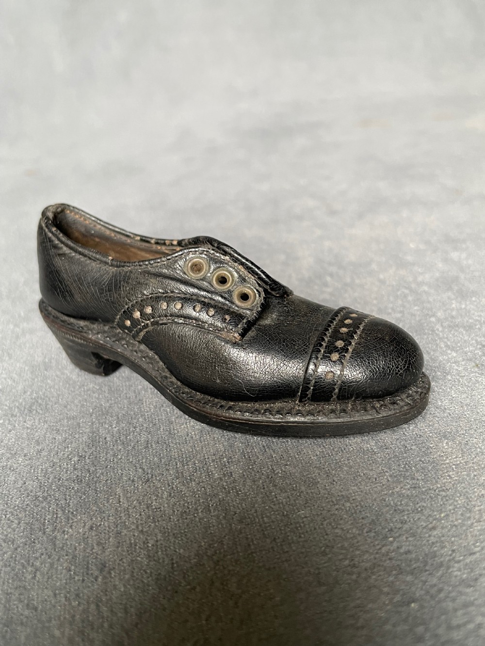A miniature black leather shoe crafted with brogue stitching and metal eyelets displayed next to a coin for scale. View VI