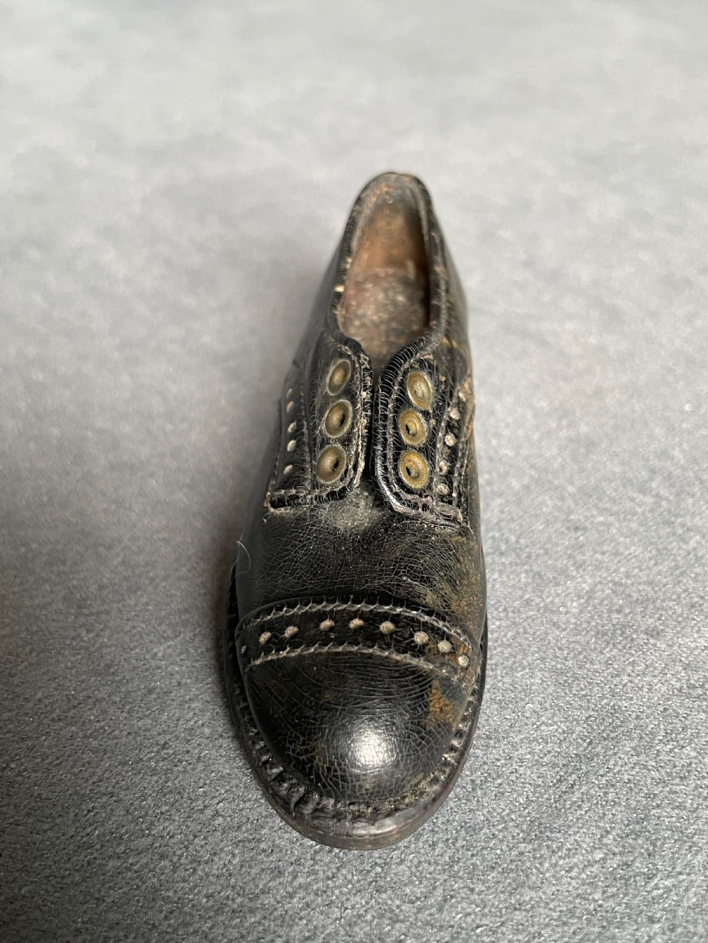 A miniature black leather shoe crafted with brogue stitching and metal eyelets displayed next to a coin for scale. View VII