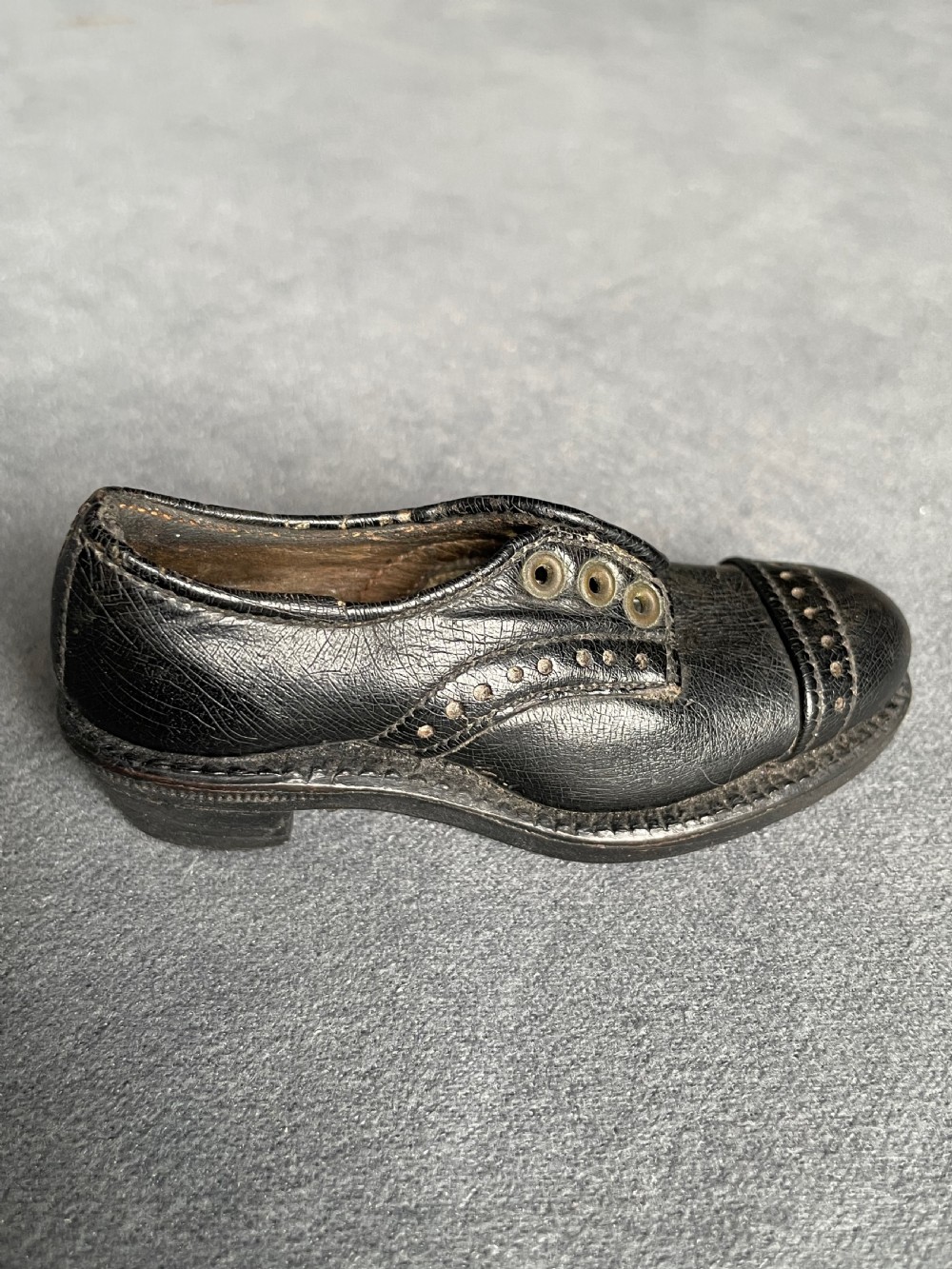 A miniature black leather shoe crafted with brogue stitching and metal eyelets displayed next to a coin for scale. View VIII