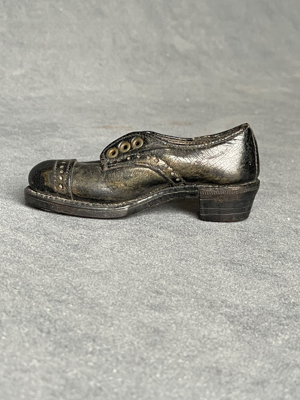 A miniature black leather shoe crafted with brogue stitching and metal eyelets displayed next to a coin for scale. View X