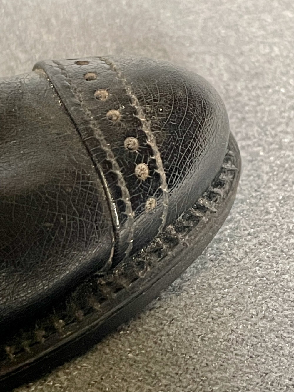 A miniature black leather shoe crafted with brogue stitching and metal eyelets displayed next to a coin for scale. View XII