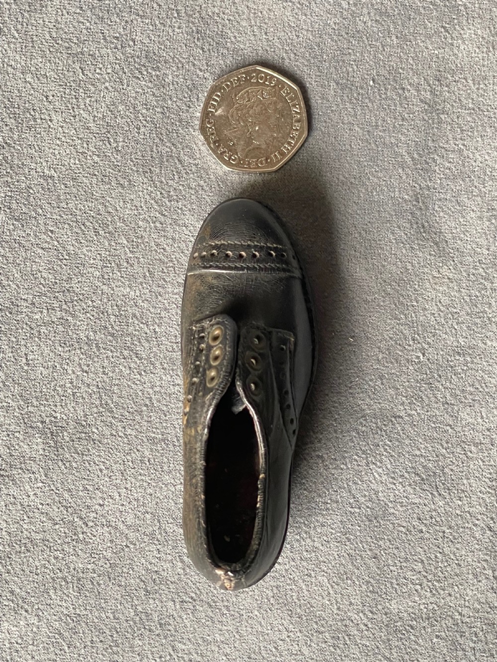 A miniature black leather shoe crafted with brogue stitching and metal eyelets displayed next to a coin for scale. View XVII