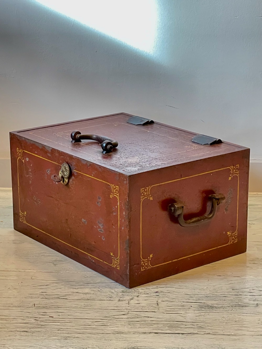 A rectangular terracotta red painted iron strongbox with gilt corner borders, top handle, side carrying handles, and a central brass keyhole escutcheon.
