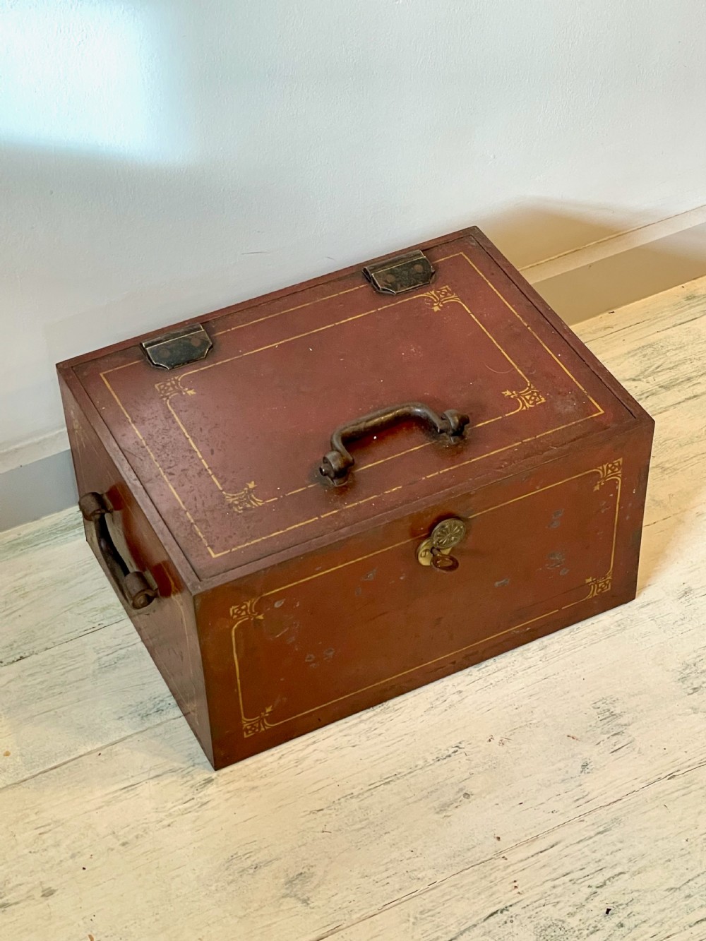 A rectangular terracotta red painted iron strongbox with gilt corner borders, top handle, side carrying handles, and a central brass keyhole escutcheon. View III