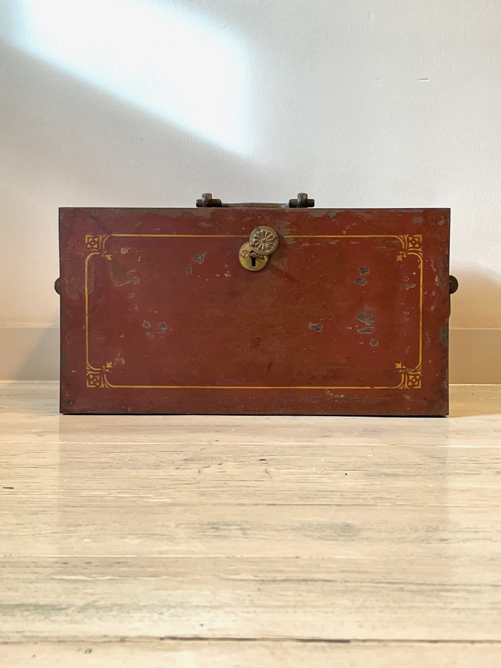 A rectangular terracotta red painted iron strongbox with gilt corner borders, top handle, side carrying handles, and a central brass keyhole escutcheon. View V