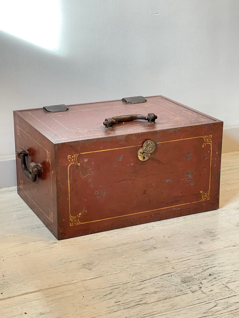 A rectangular terracotta red painted iron strongbox with gilt corner borders, top handle, side carrying handles, and a central brass keyhole escutcheon. View VI