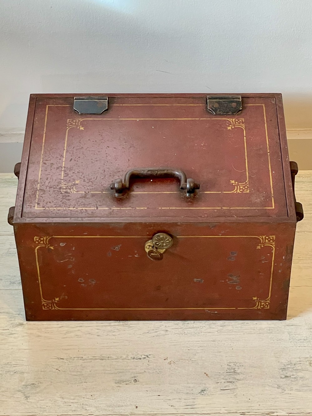A rectangular terracotta red painted iron strongbox with gilt corner borders, top handle, side carrying handles, and a central brass keyhole escutcheon. View VII