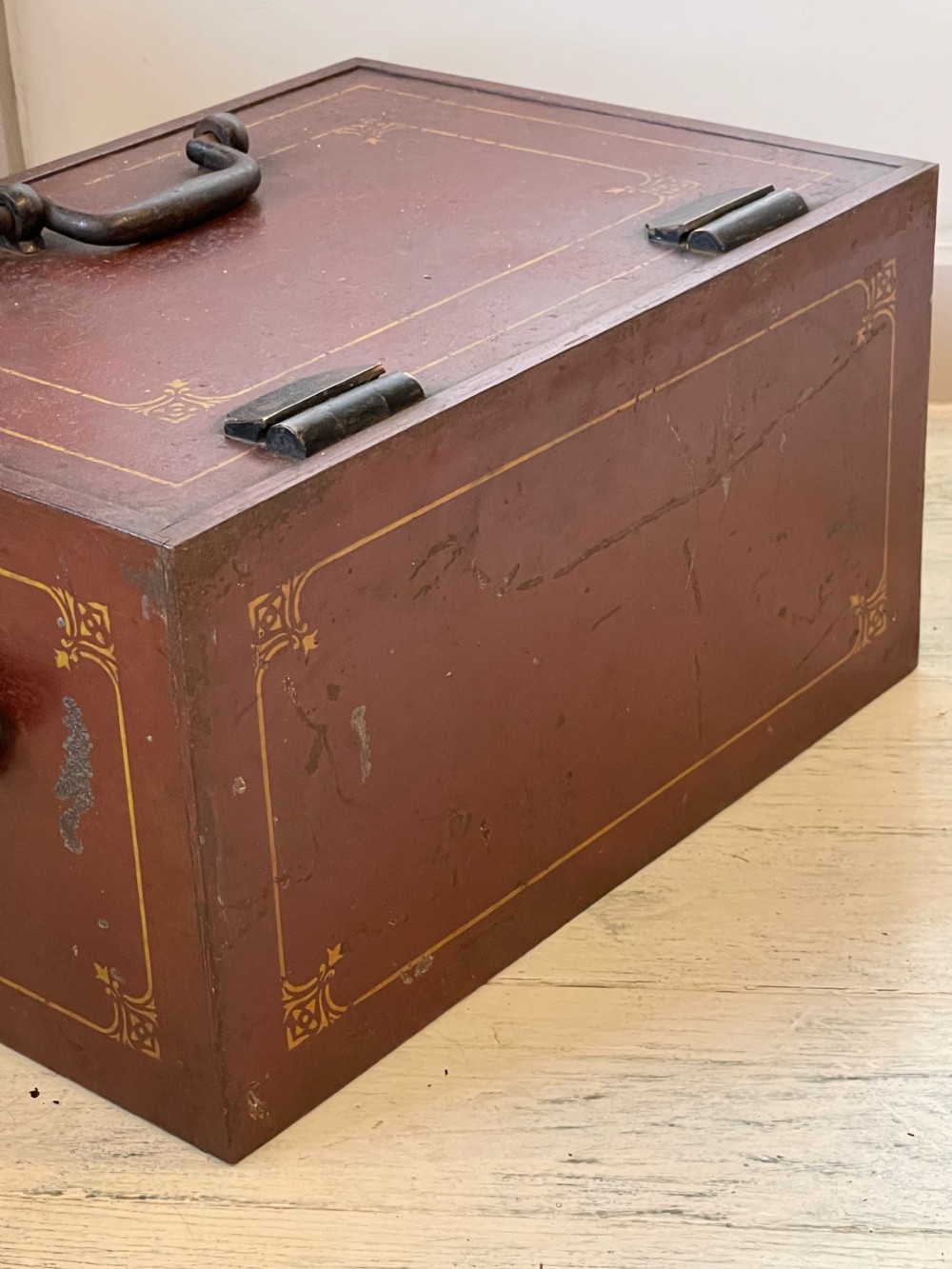 A rectangular terracotta red painted iron strongbox with gilt corner borders, top handle, side carrying handles, and a central brass keyhole escutcheon. View X