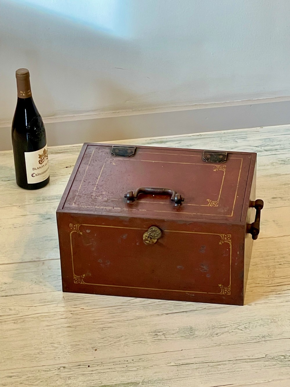 A rectangular terracotta red painted iron strongbox with gilt corner borders, top handle, side carrying handles, and a central brass keyhole escutcheon. View XIII