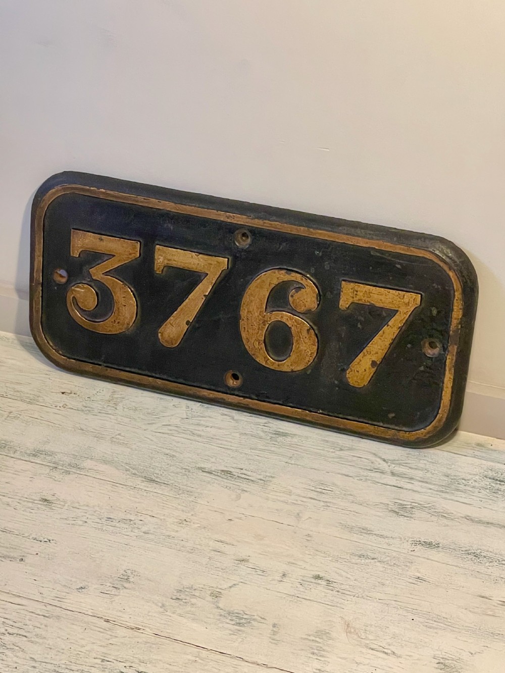 A rectangular cast metal locomotive number plate featuring raised digits 3767 in gold tone against a black background. View III