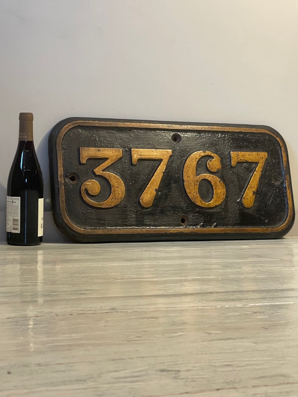 A rectangular cast metal locomotive number plate featuring raised digits 3767 in gold tone against a black background. View V