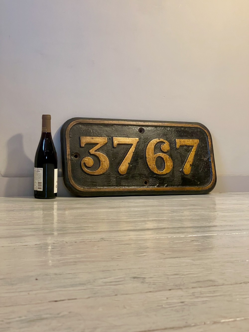 A rectangular cast metal locomotive number plate featuring raised digits 3767 in gold tone against a black background. View VI