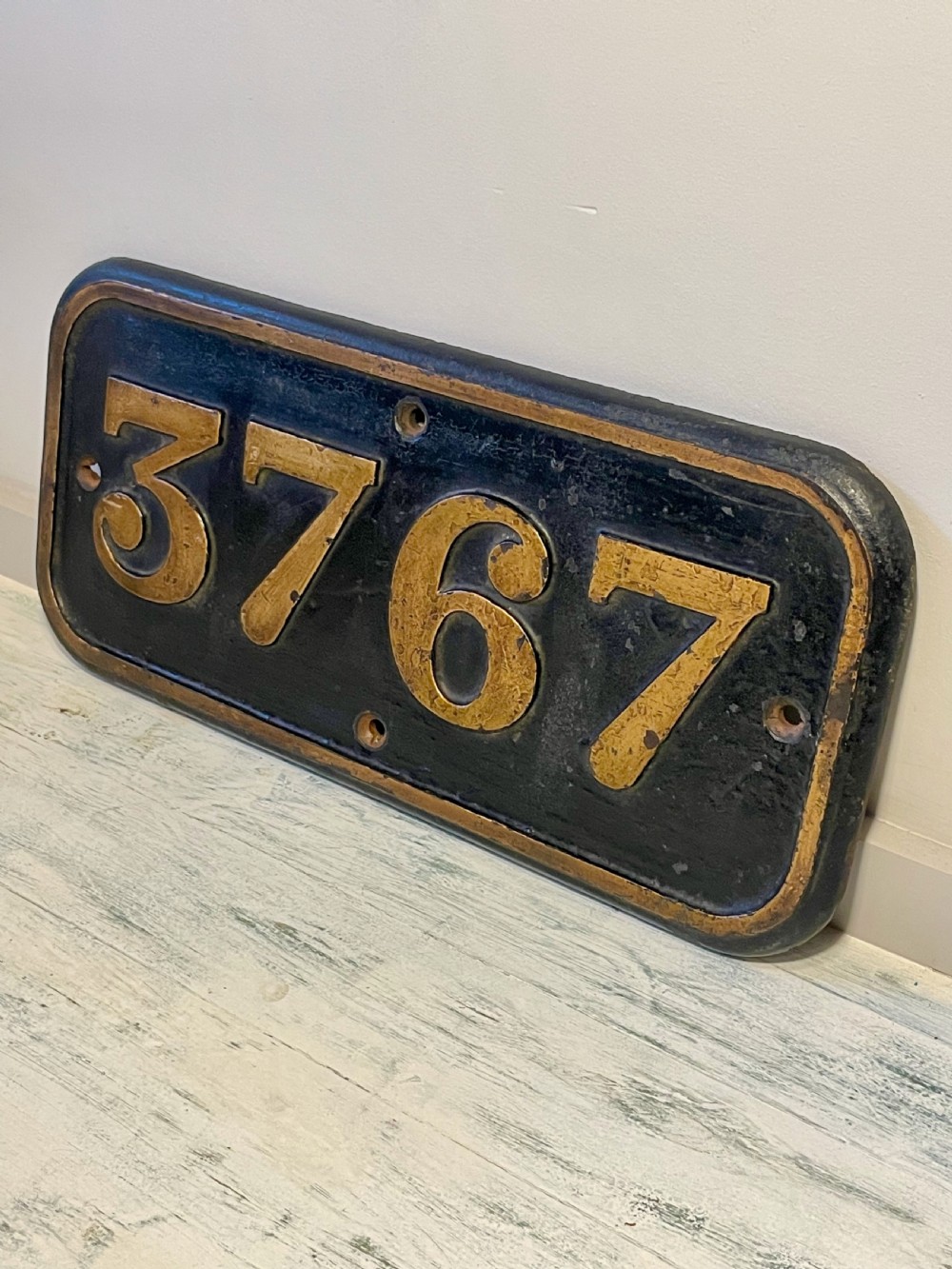 A rectangular cast metal locomotive number plate featuring raised digits 3767 in gold tone against a black background. View IX