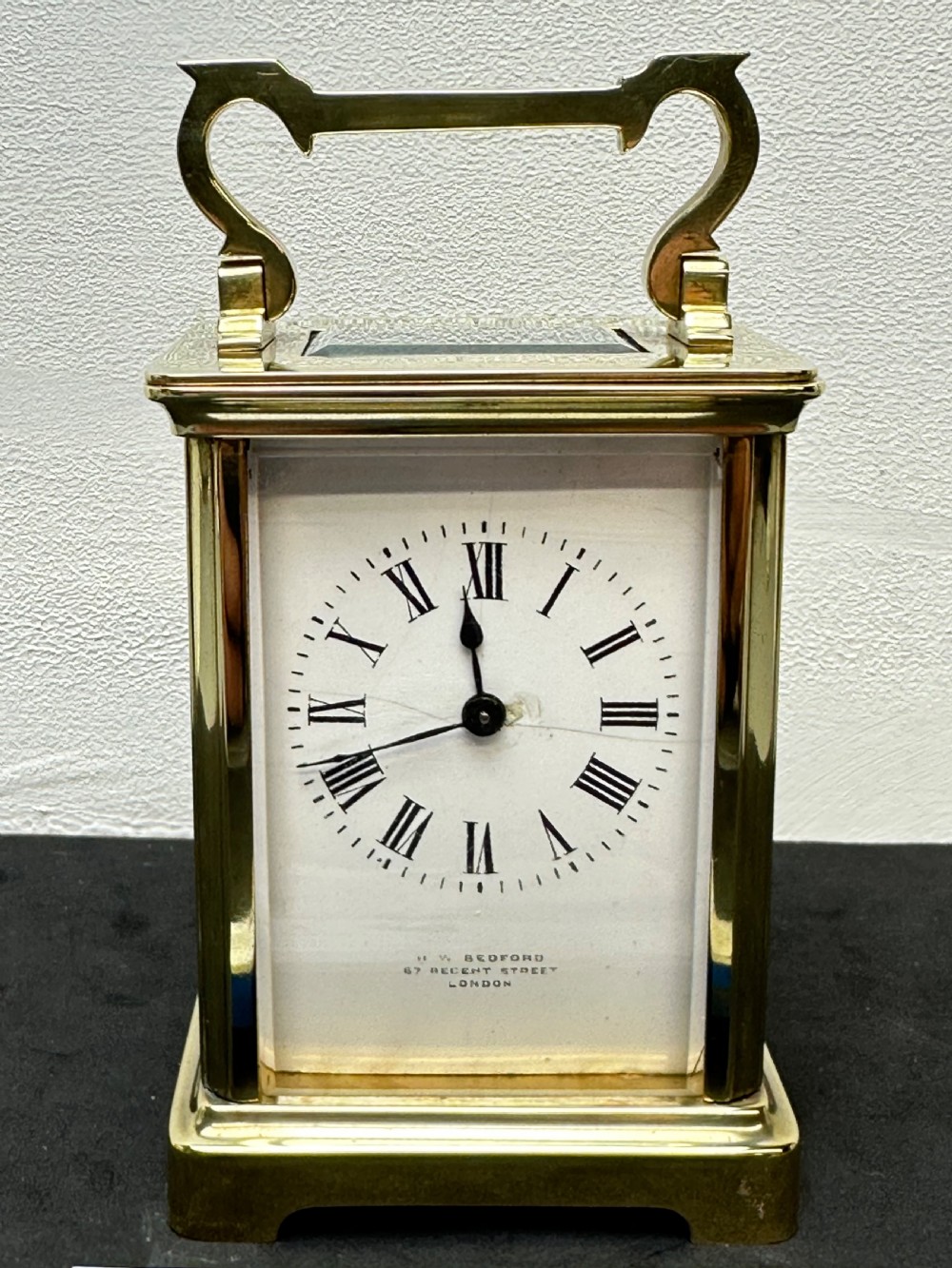 A rectangular brass French carriage clock with clear glass panels, a top carrying handle, and a white enamel dial with black Roman numerals. View II