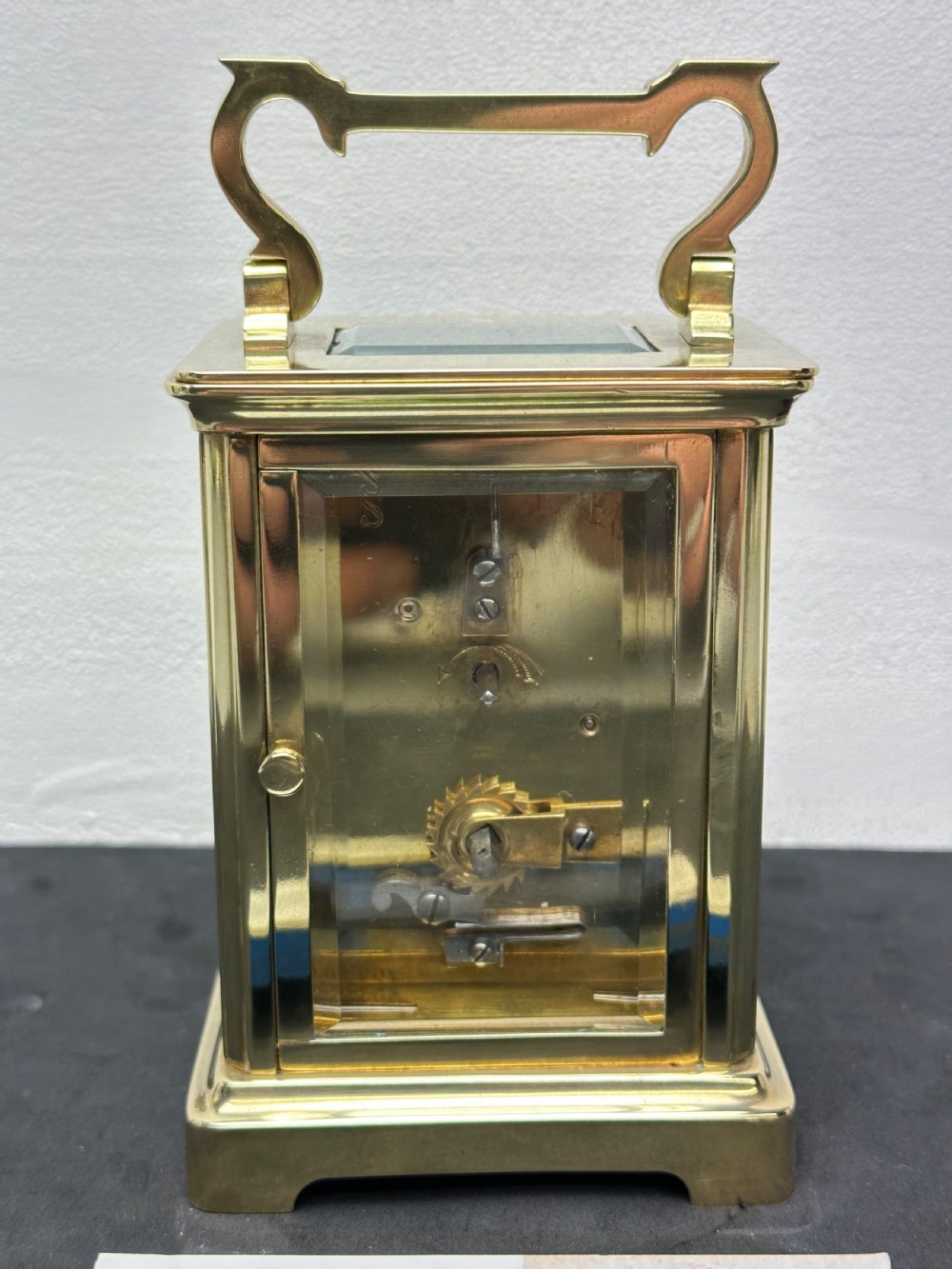 A rectangular brass French carriage clock with clear glass panels, a top carrying handle, and a white enamel dial with black Roman numerals. View IV