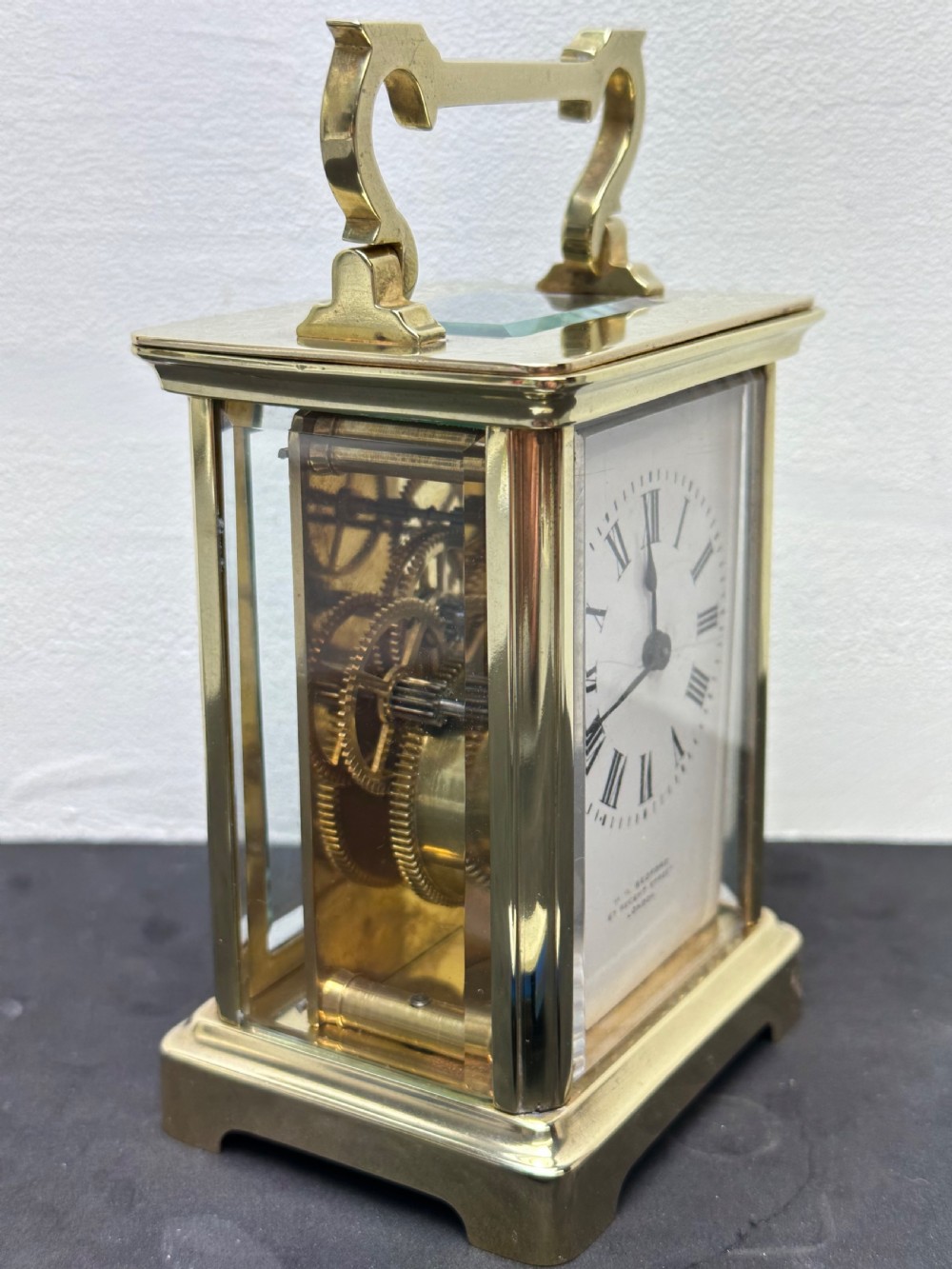 A rectangular brass French carriage clock with clear glass panels, a top carrying handle, and a white enamel dial with black Roman numerals. View VI