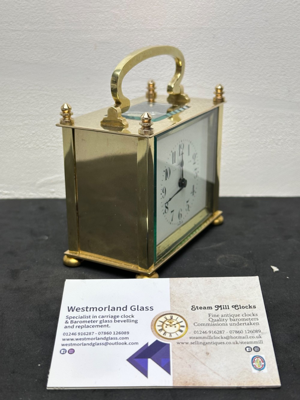 A small rectangular brass carriage clock featuring a top handle, decorative finials, and a white dial with black Arabic numerals. View II