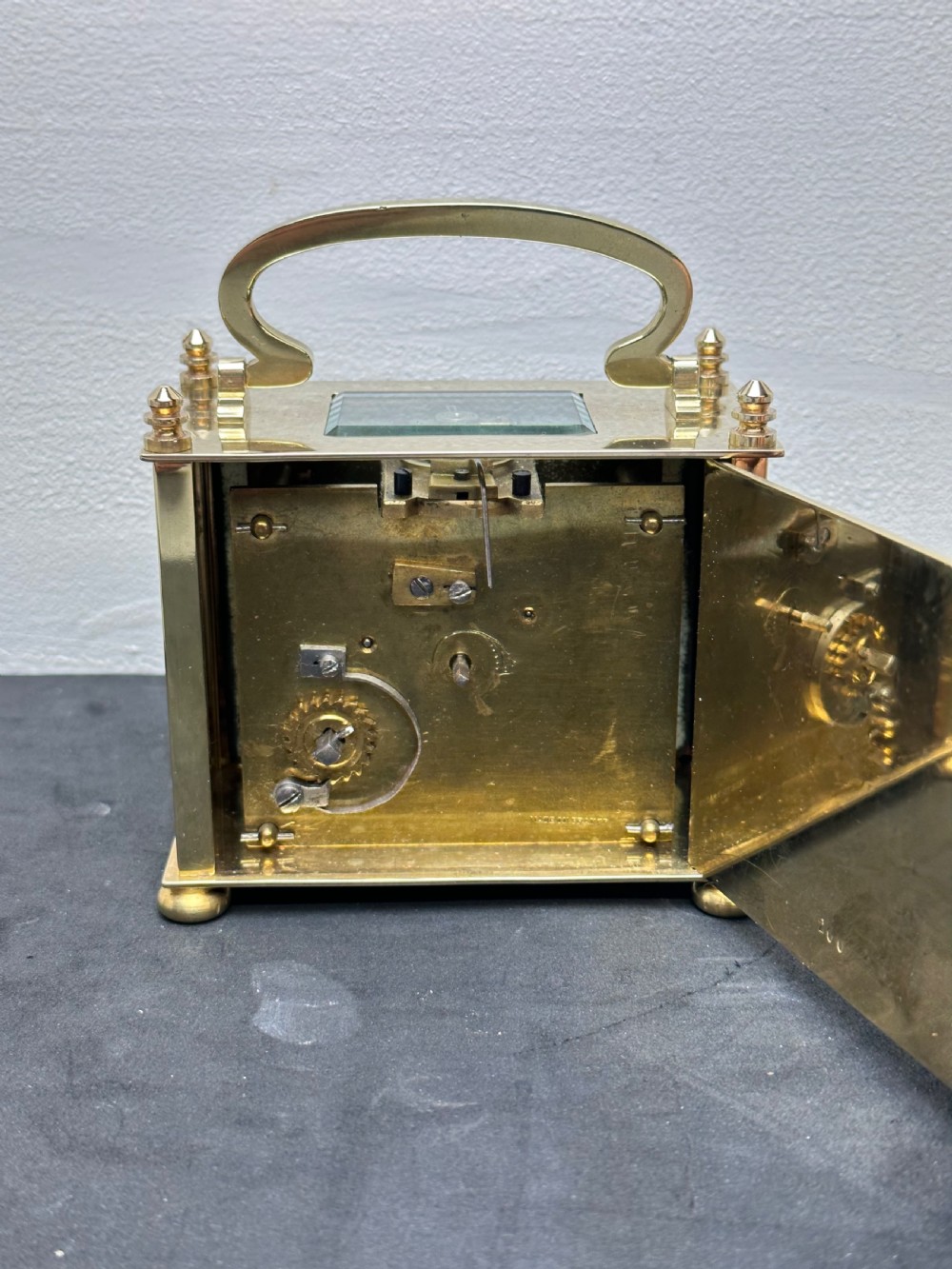 A small rectangular brass carriage clock featuring a top handle, decorative finials, and a white dial with black Arabic numerals. View III
