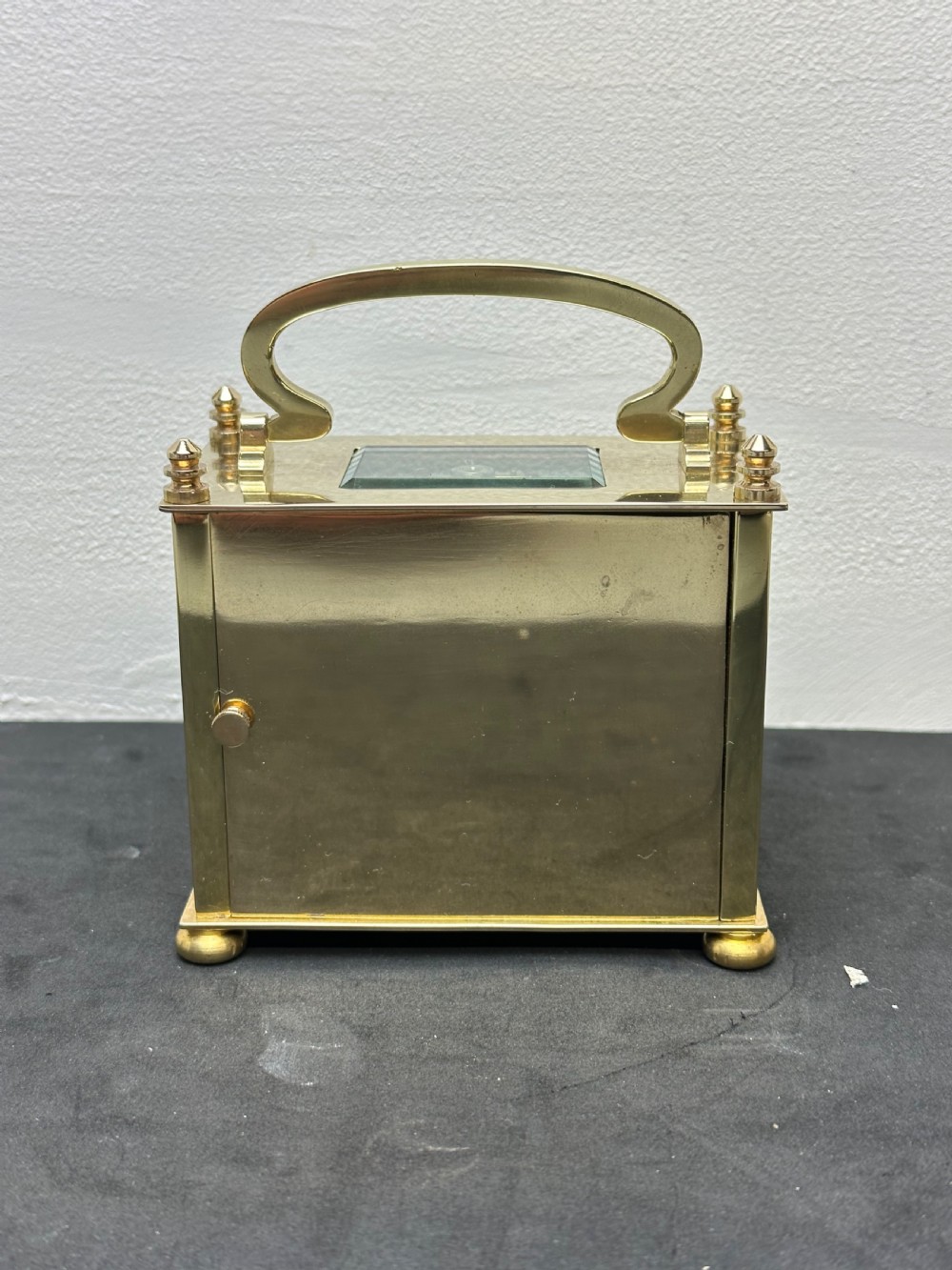 A small rectangular brass carriage clock featuring a top handle, decorative finials, and a white dial with black Arabic numerals. View IV
