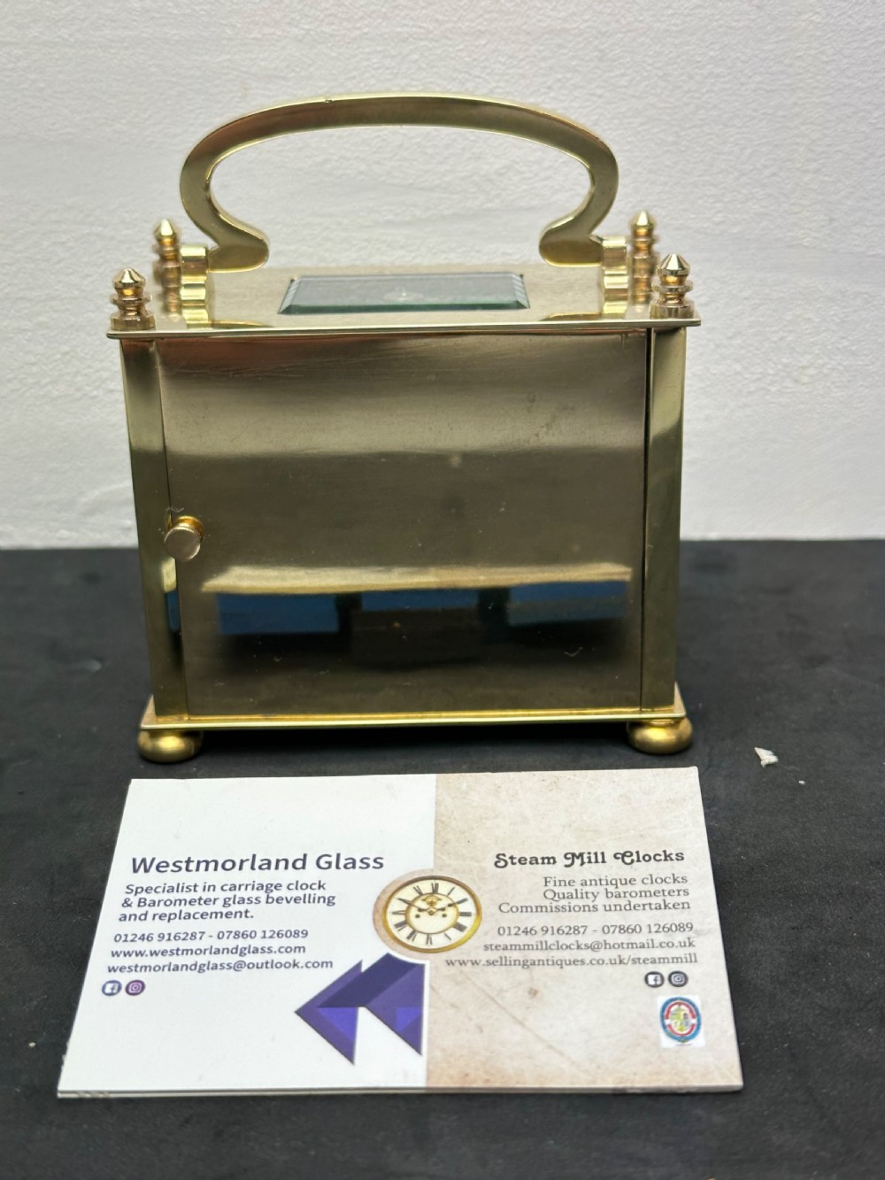 A small rectangular brass carriage clock featuring a top handle, decorative finials, and a white dial with black Arabic numerals. View V