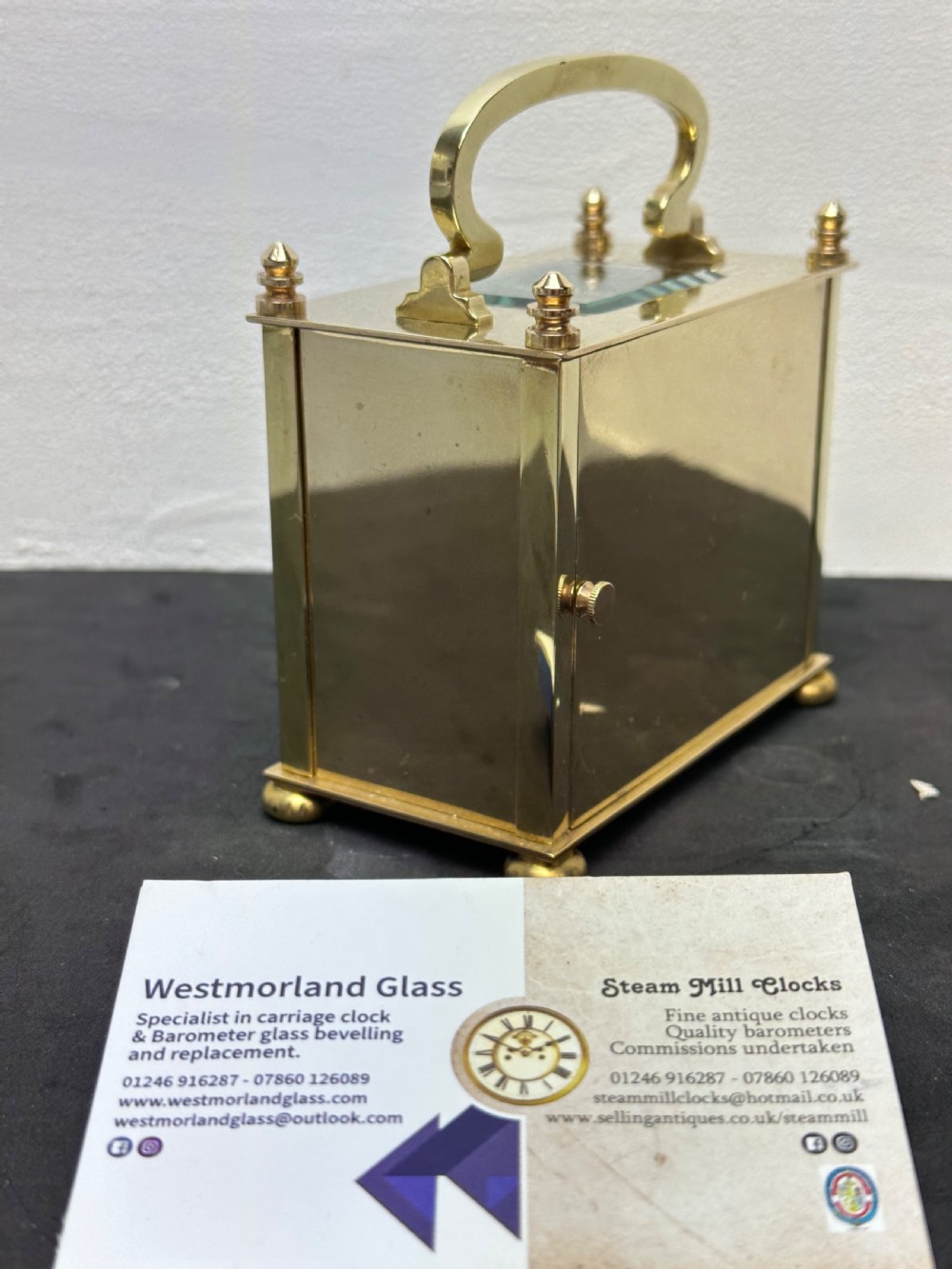 A small rectangular brass carriage clock featuring a top handle, decorative finials, and a white dial with black Arabic numerals. View VI