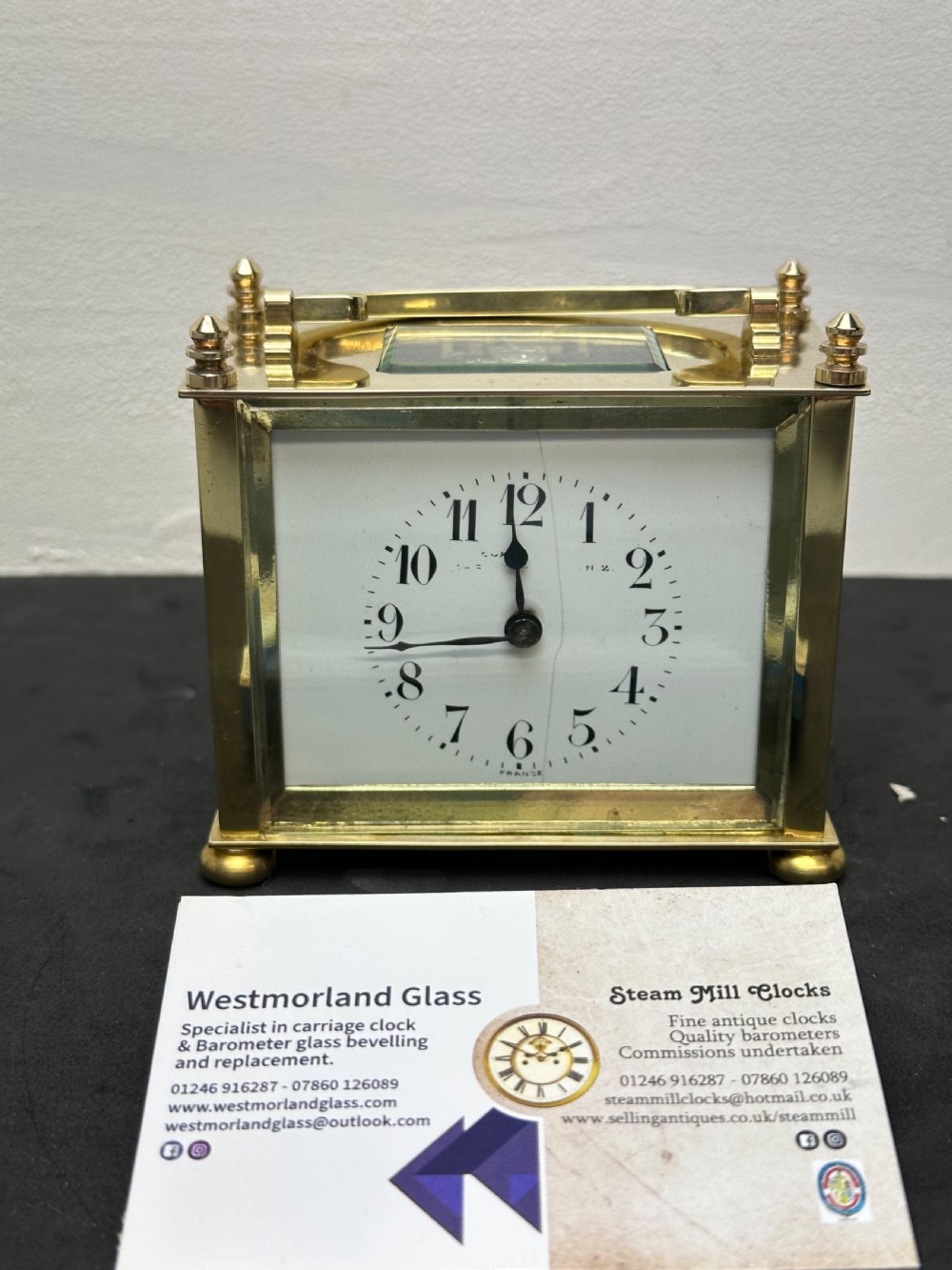 A small rectangular brass carriage clock featuring a top handle, decorative finials, and a white dial with black Arabic numerals. View VII