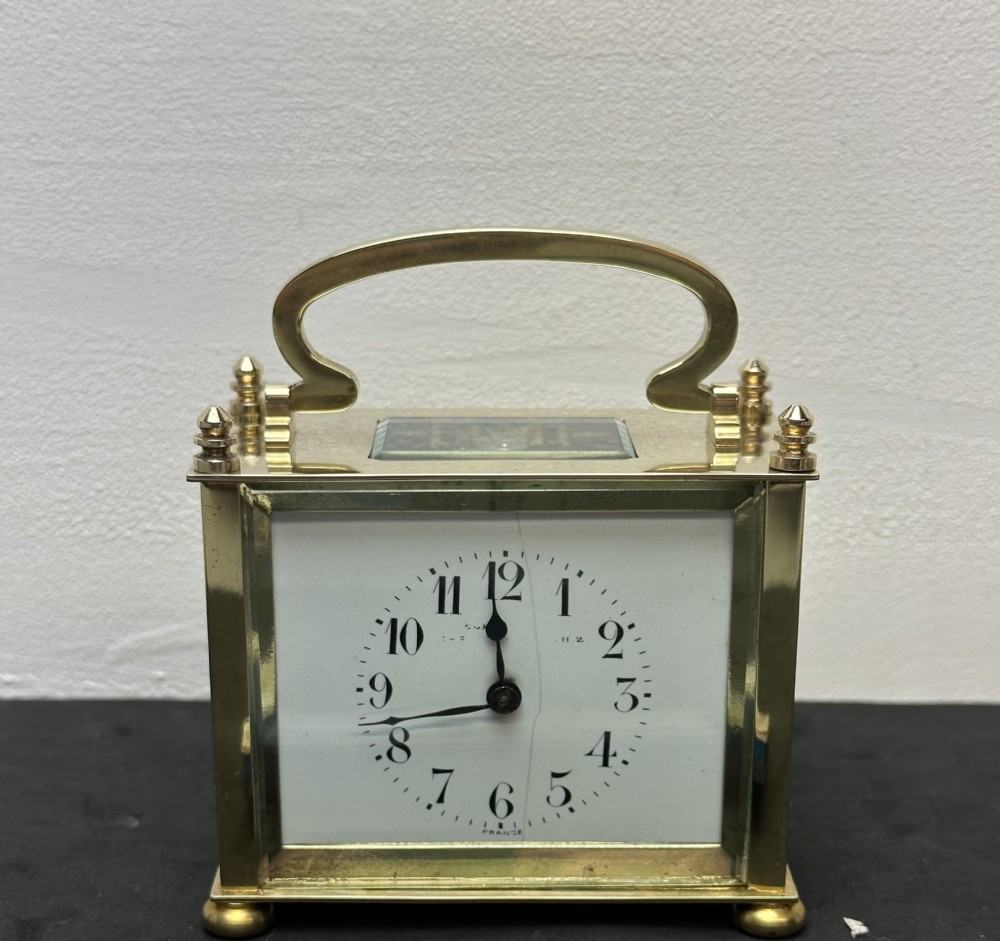 A small rectangular brass carriage clock featuring a top handle, decorative finials, and a white dial with black Arabic numerals.