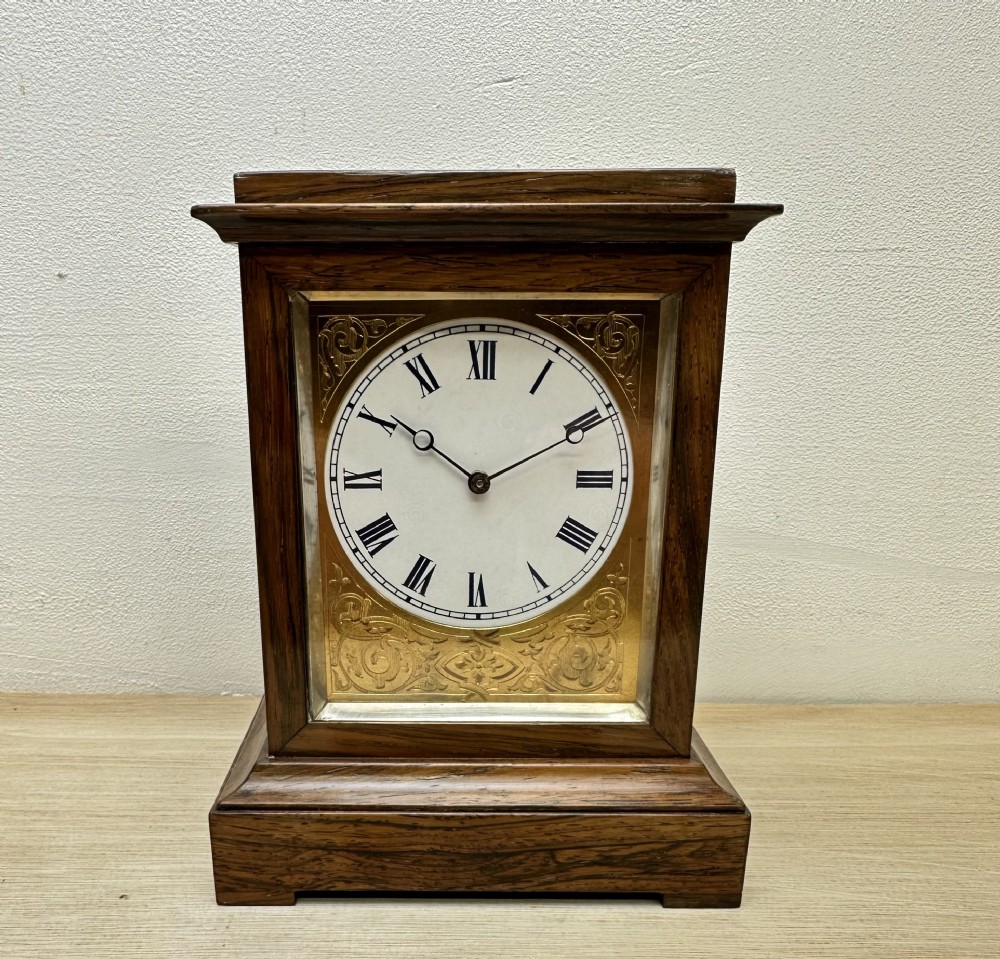 A rectangular dark rosewood mantel clock featuring an enamel dial with Roman numerals surrounded by decorative brass spandrels.