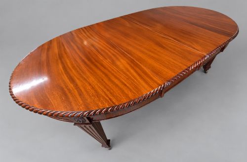 fig 1. An oval mahogany extending dining table with a carved edge and winding mechanism, surrounded by eight carved wooden chairs featuring blue floral needlepoint seats.