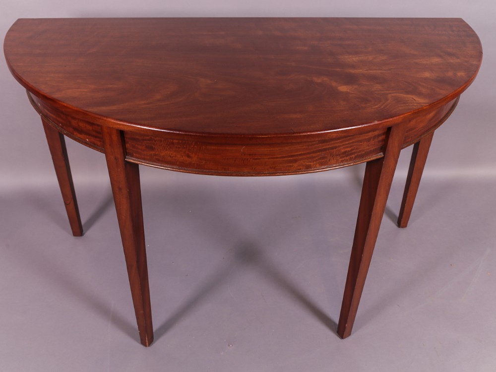A Georgian mahogany semi-circular D-end console table featuring a figured wood top and four square tapered legs.