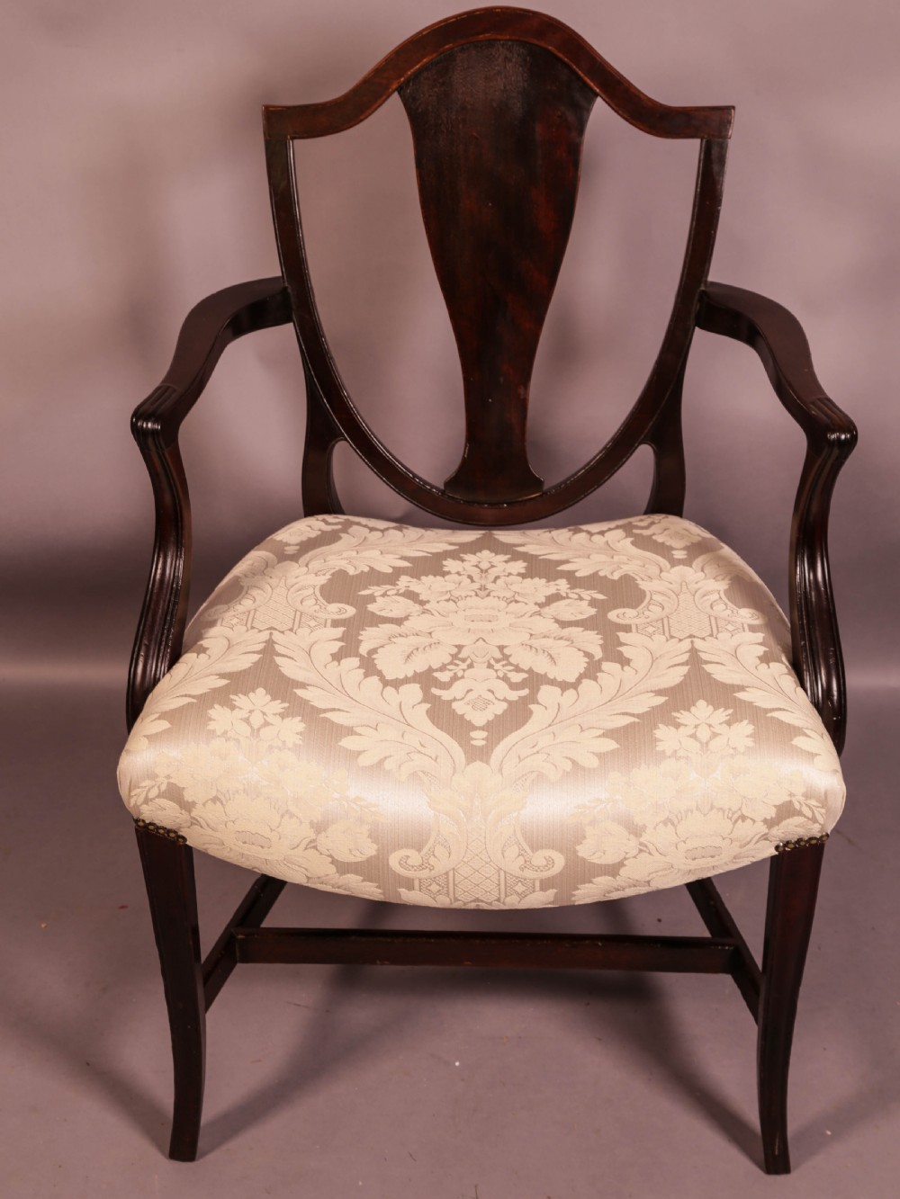 A dark mahogany Hepplewhite period carver armchair featuring a carved open backrest, curved arms, and a cream patterned damask seat.